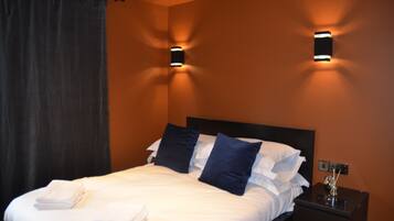 A neatly made bed with white linens and two dark blue pillows, a black nightstand with a lamp, and two wall-mounted sconces on an orange wall.