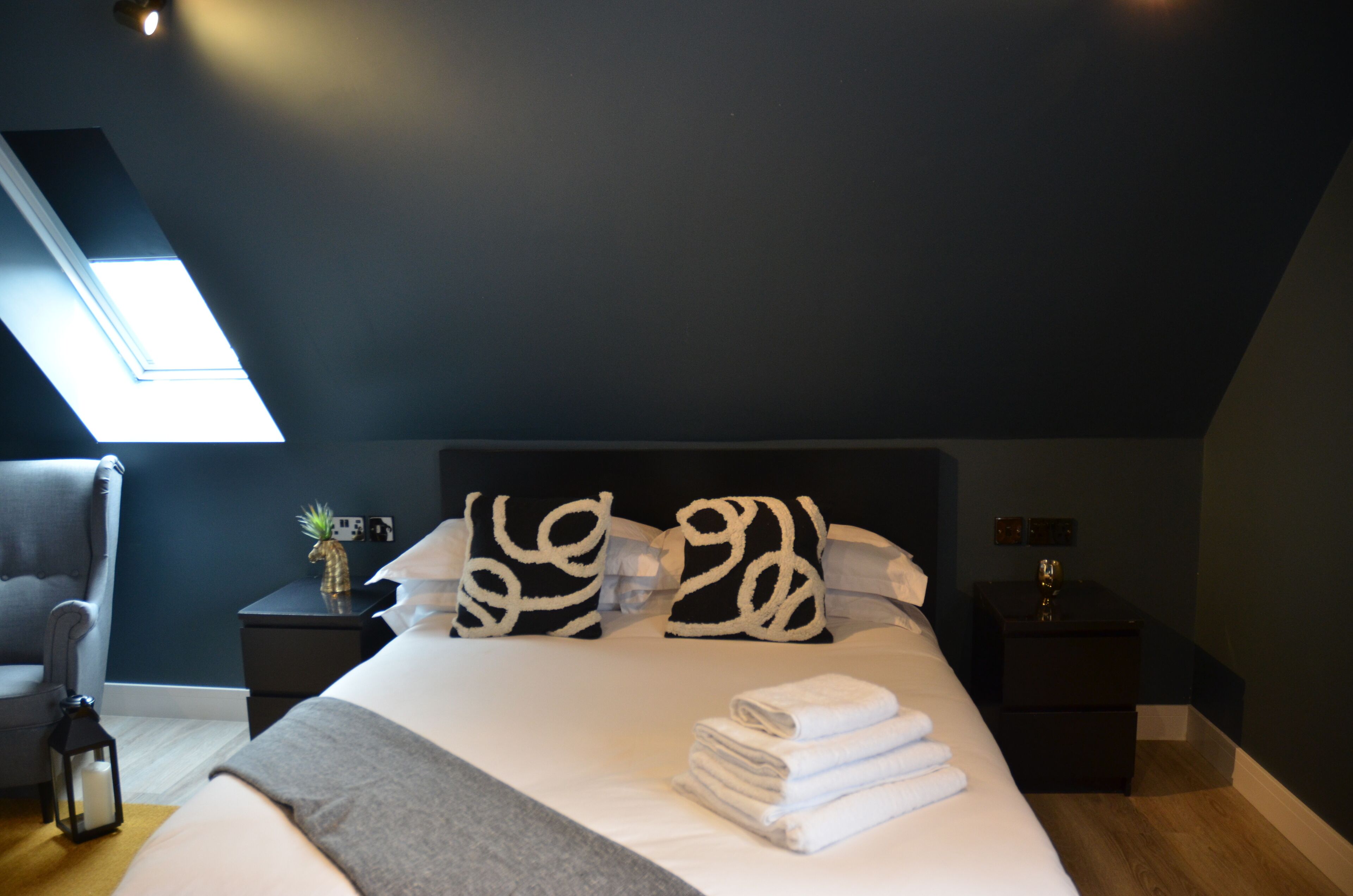 Een net opgemaakt bed met witte lakens en decoratieve kussens, een klein nachtkastje met een plant en een kaarsje, en een donkerblauwe wand met een dakraam.