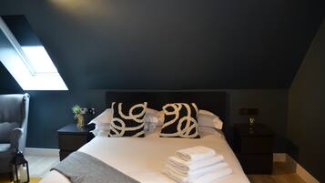 Een net opgemaakt bed met witte lakens en decoratieve kussens, een klein nachtkastje met een plant en een kaarsje, en een donkerblauwe wand met een dakraam.