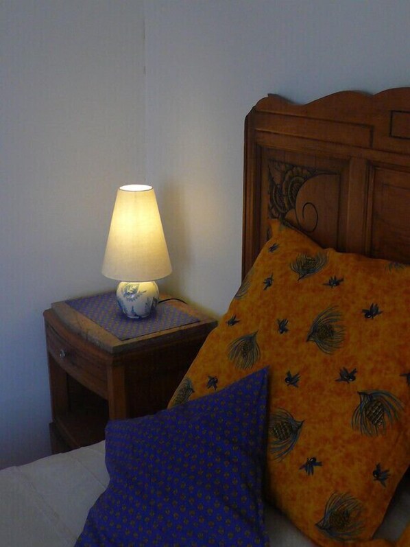 A bed with a blue pillow and an orange pillow with a leaf pattern, a nightstand with a lamp, and a wooden headboard. - Le Cadran Solaire (Cabrières)
