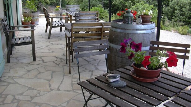 Un patio al aire libre con bancos y mesas de madera, rodeado de plantas en macetas y un barril grande.