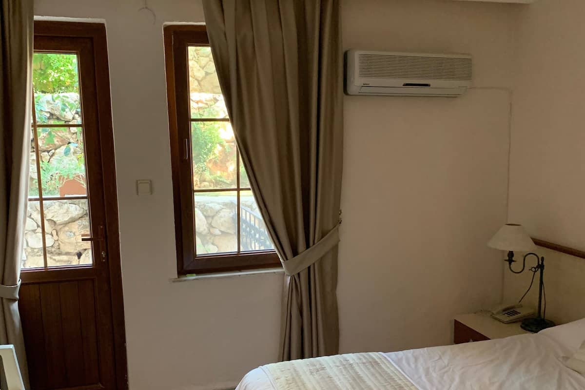 A bedroom with a bed, a nightstand with a lamp, a window with curtains, and an air conditioning unit.