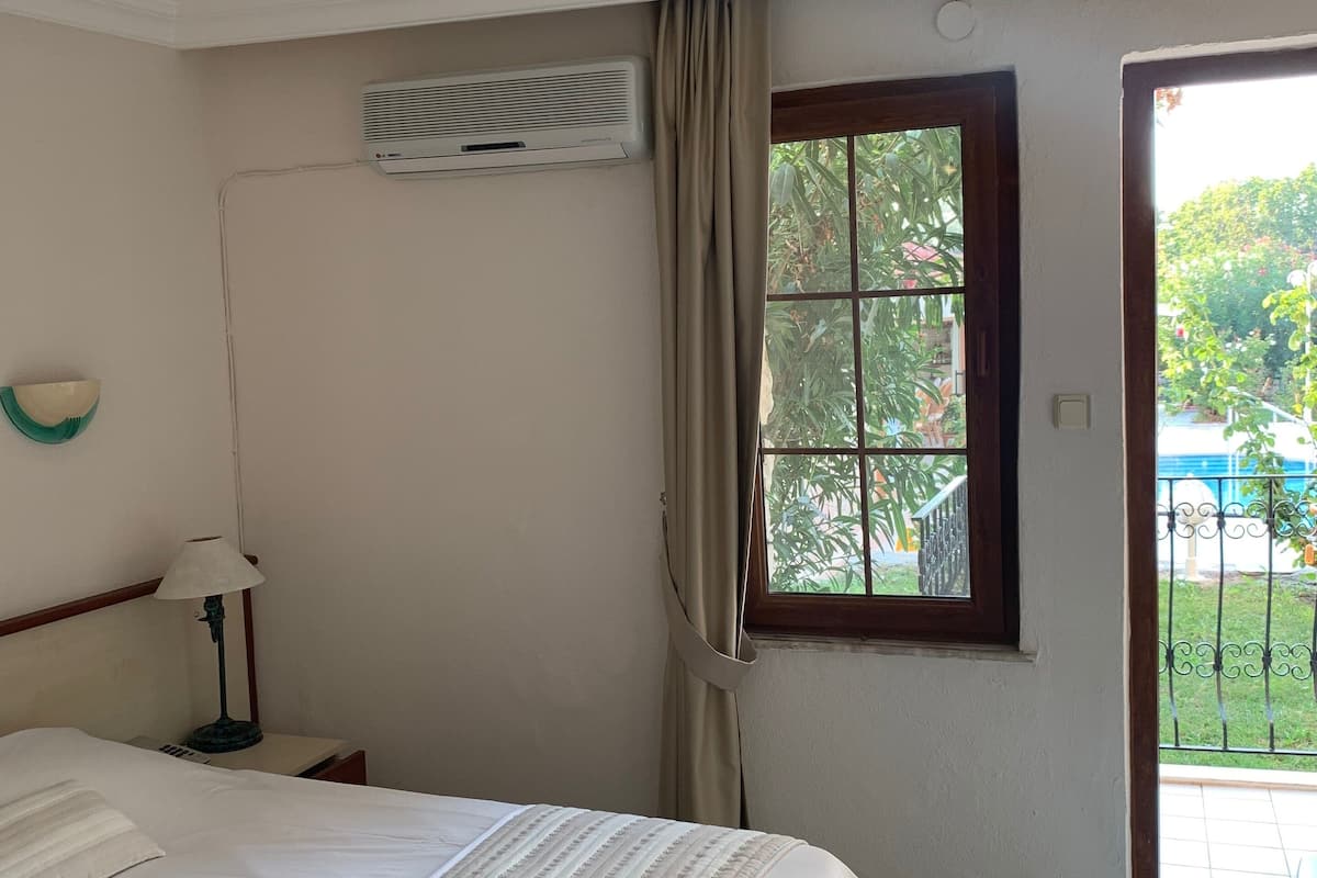 A bedroom with a bed, an air conditioning unit, a window with curtains, and a view of the outside.