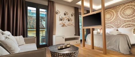A modern hotel room with a sofa, a round table with decorative items, a bed with a patterned headboard, and a TV mounted on a wooden frame.