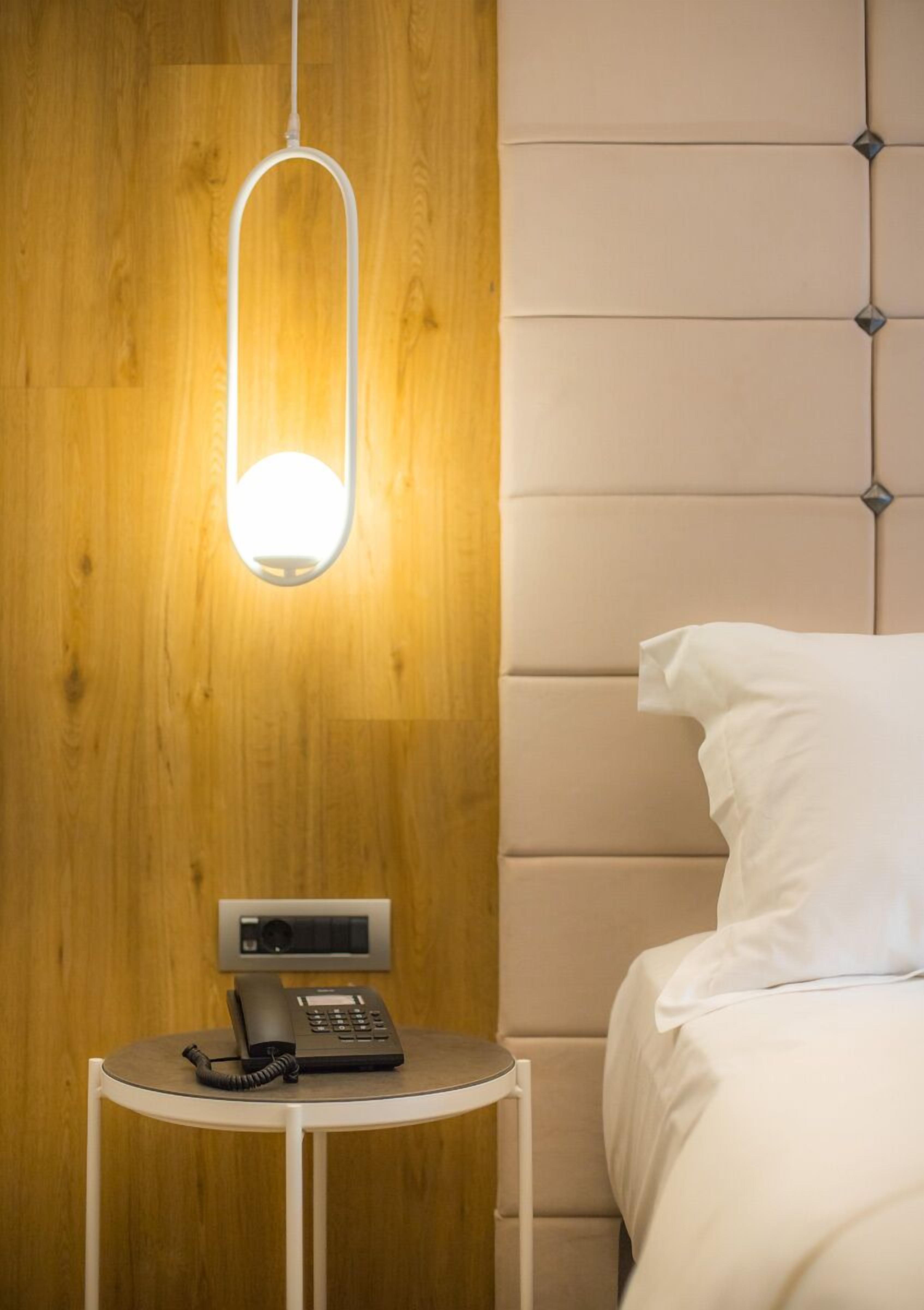 A hotel room with a wooden panel wall, a modern hanging light fixture, a bedside table with a telephone, and a bed with white bedding.