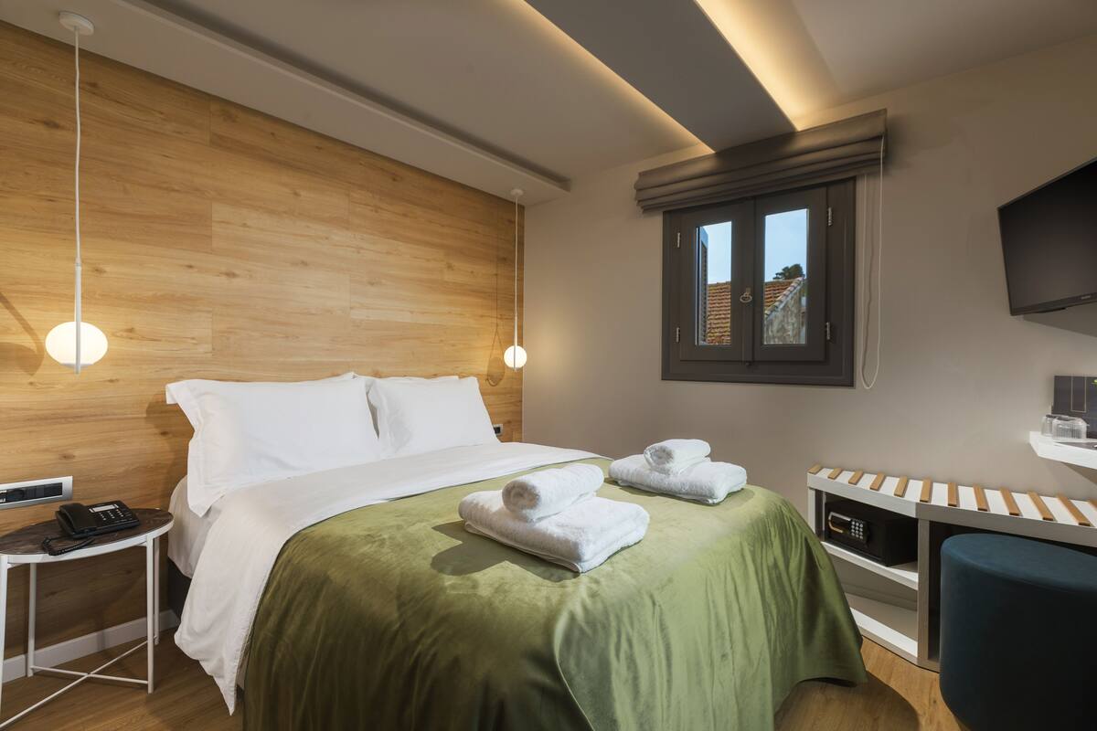 Una habitación de hotel moderna con cabecera de madera, cama con ropa de cama verde, dos toallas blancas, una mesita de noche con un teléfono, un escritorio pequeño con una silla y un televisor montado en la pared.