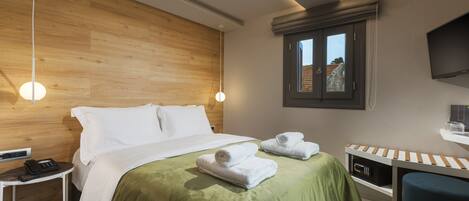 A modern hotel room with a wooden headboard, a bed with green bedding, two white towels, a bedside table with a phone, a small desk with a chair, and a TV mounted on the wall.