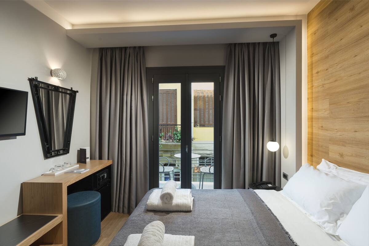 Habitación de hotel con cama, escritorio, silla, televisión y balcón con cortinas.