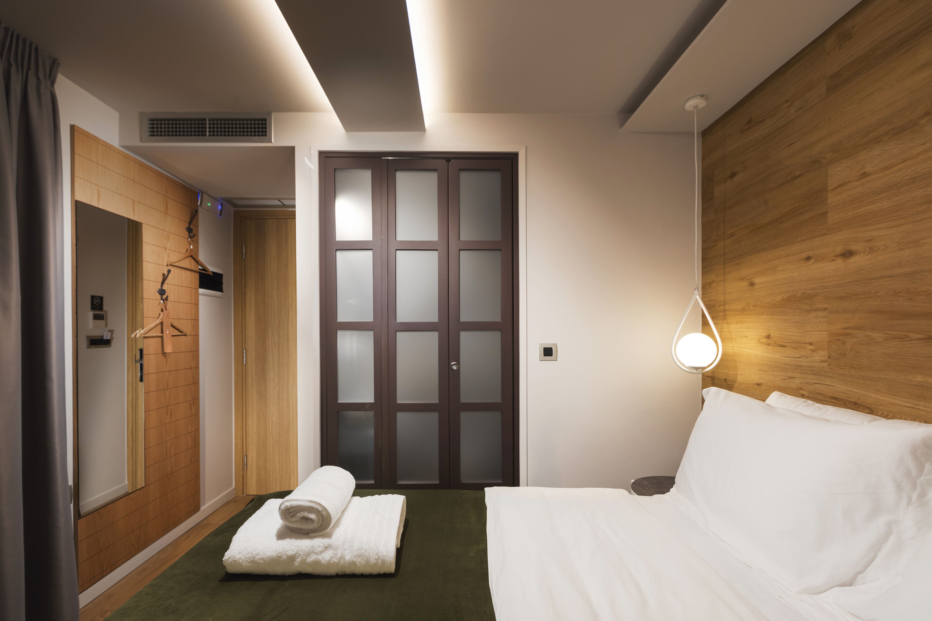 A hotel room with a bed, a wooden wardrobe, a towel, a hanging coat rack, and a wall-mounted light fixture.
