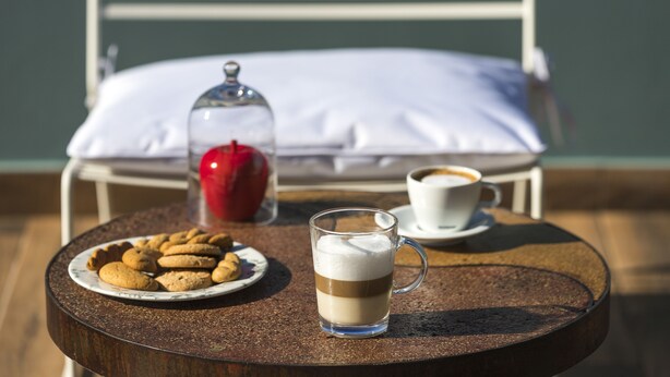 Une table sur laquelle se trouvent une assiette de biscuits, un verre contenant une boisson à plusieurs couches, une tasse de café et une bougie rouge. La table est ronde et une chaise se trouve à l’arrière-plan.