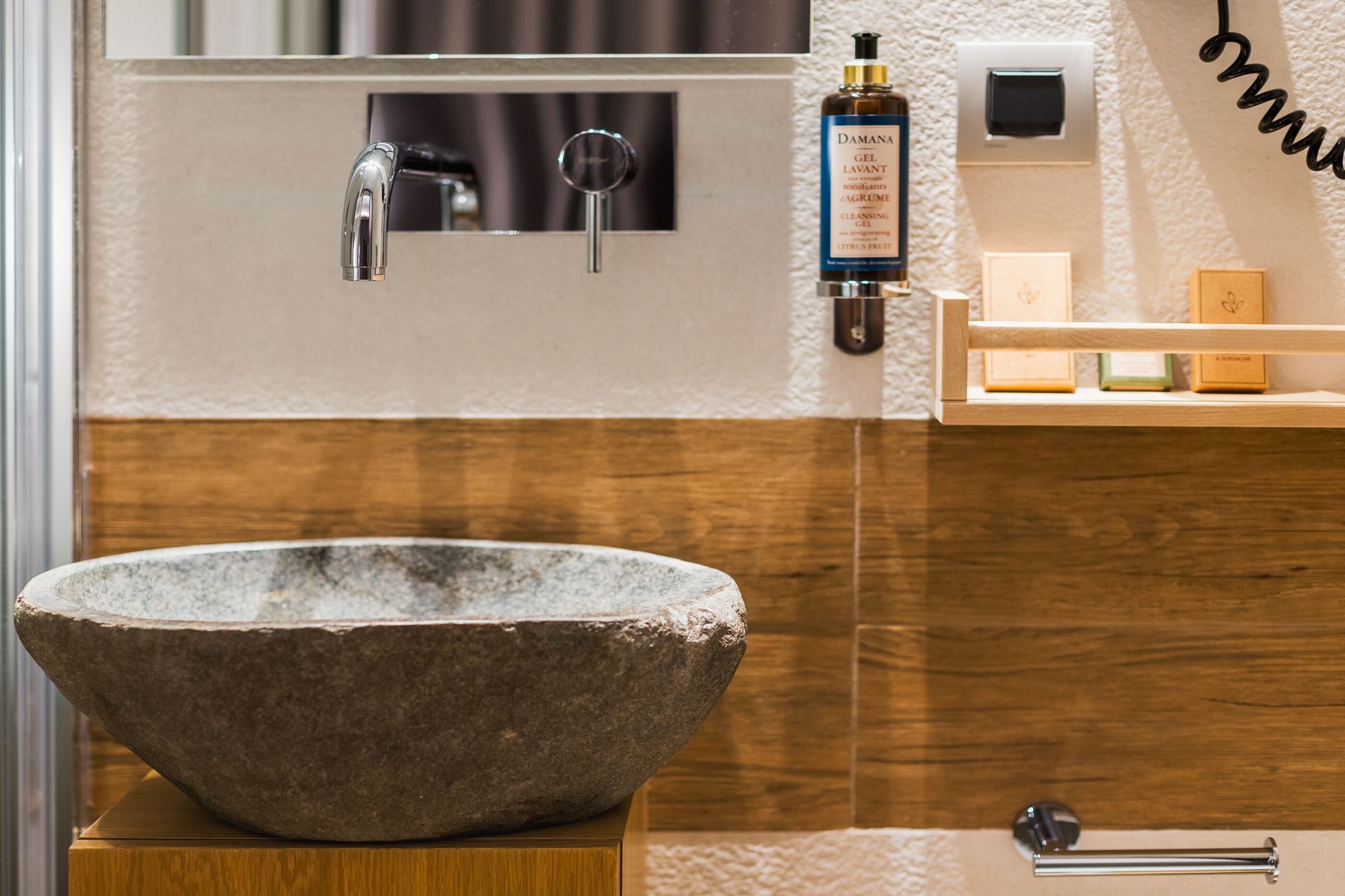 Une salle de bain avec un lavabo en pierre, un robinet moderne, un flacon de gel de Damana et un distributeur de savon sur une étagère en bois.