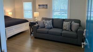 A bedroom with a bed, a gray sofa, two windows with blinds, a ceiling fan, and a nightstand with a lamp.