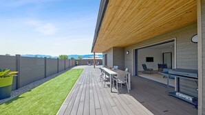Una moderna veranda esterna con un pavimento in legno, un'area pranzo con sedie e tavolo, e vista sulle montagne sullo sfondo.