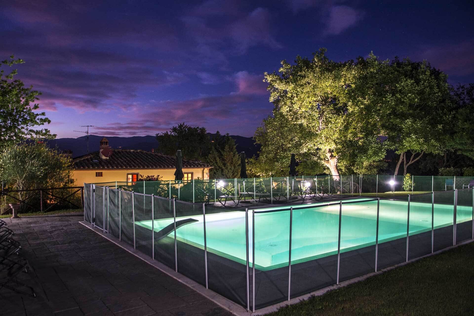 A rectangular swimming pool with a glass enclosure, surrounded by a fence and outdoor seating.