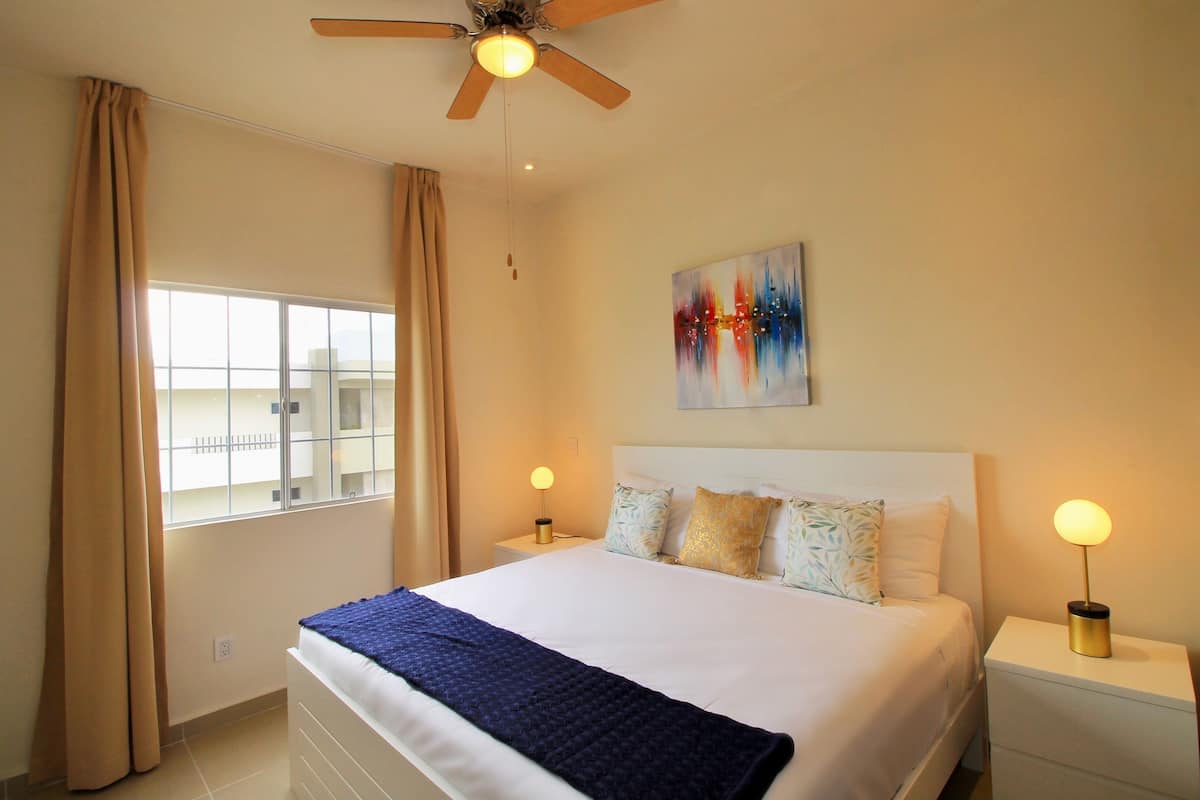 Habitación de hotel con cama, dos mesitas de noche con lámparas, ventilador de techo, ventana con cortinas y un cuadro en la pared.