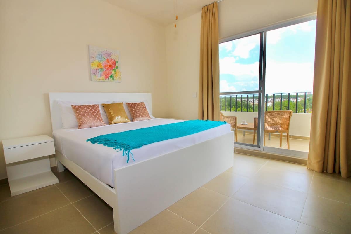 Una habitación de hotel con cama, mesita de noche, un cuadro, un balcón con una silla y una mesa.