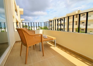 A balcony with a wicker chair and a small table, overlooking a residential area with buildings.