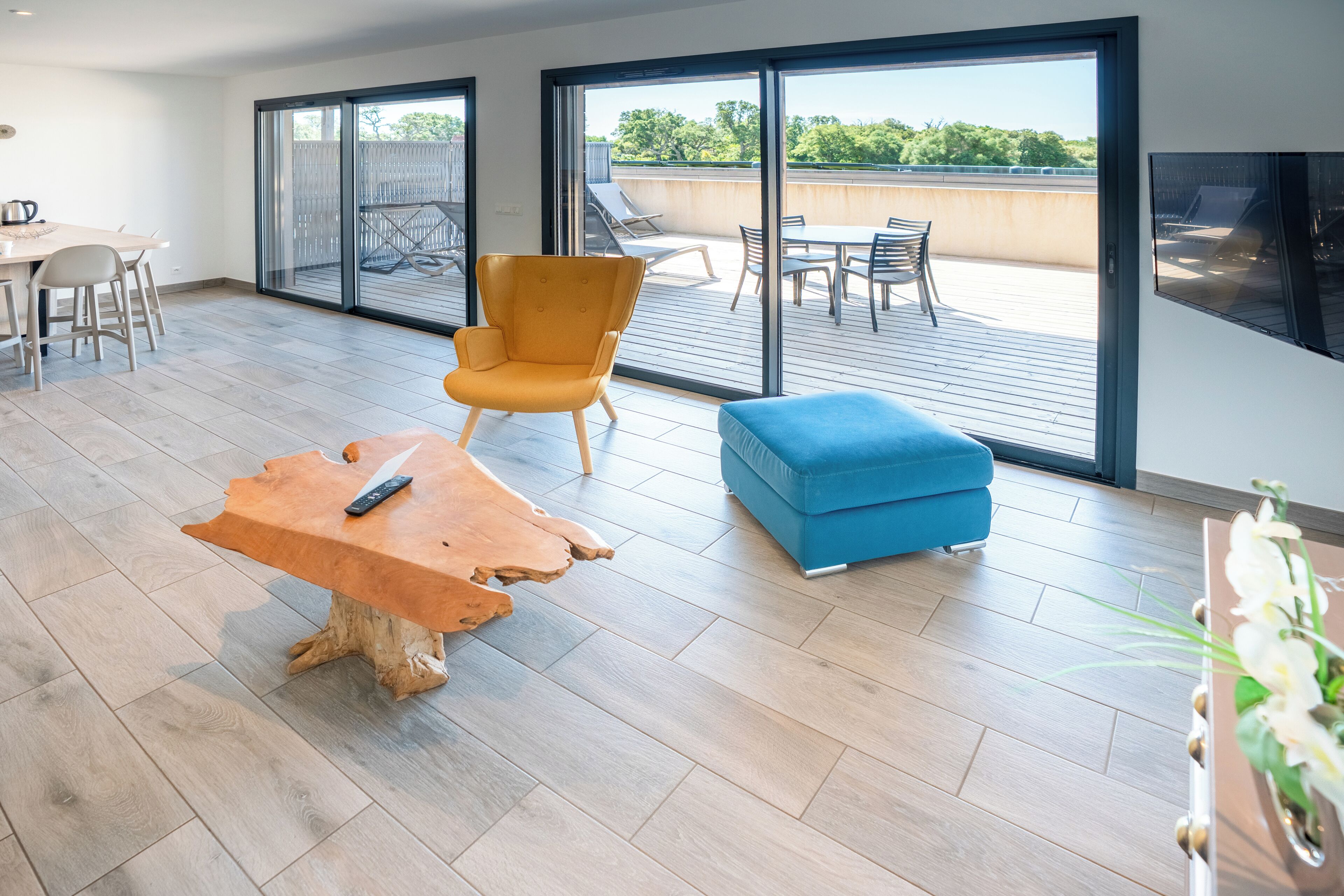 A modern living room with a wooden coffee table, a blue ottoman, a yellow armchair, and a dining area with a table and chairs.