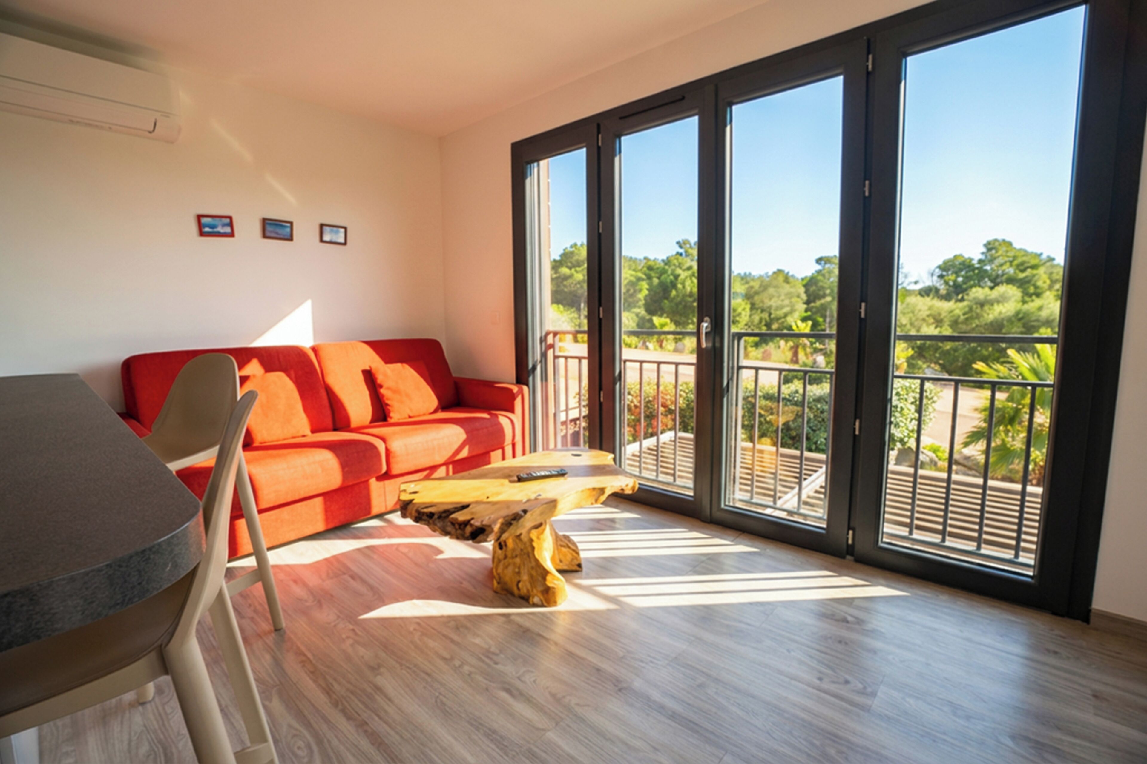 Sala de estar moderna com um sofá vermelho, uma mesa de centro de madeira e uma área de jantar com mesa e cadeiras. Grandes portas de vidro dão acesso a uma varanda com vista para o verde.
