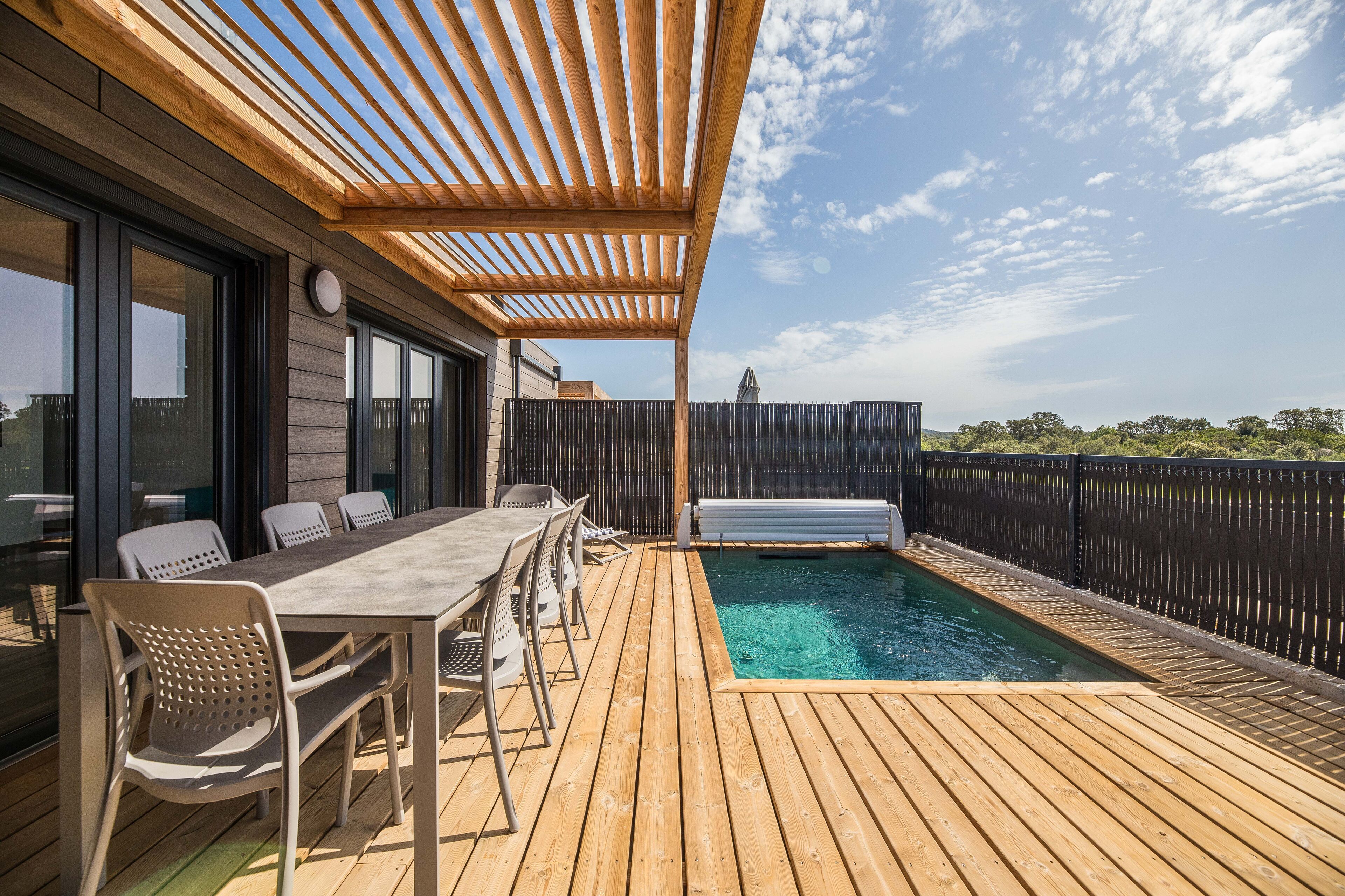 Een houten terras met zwembad, buitenmeubels en een pergola.