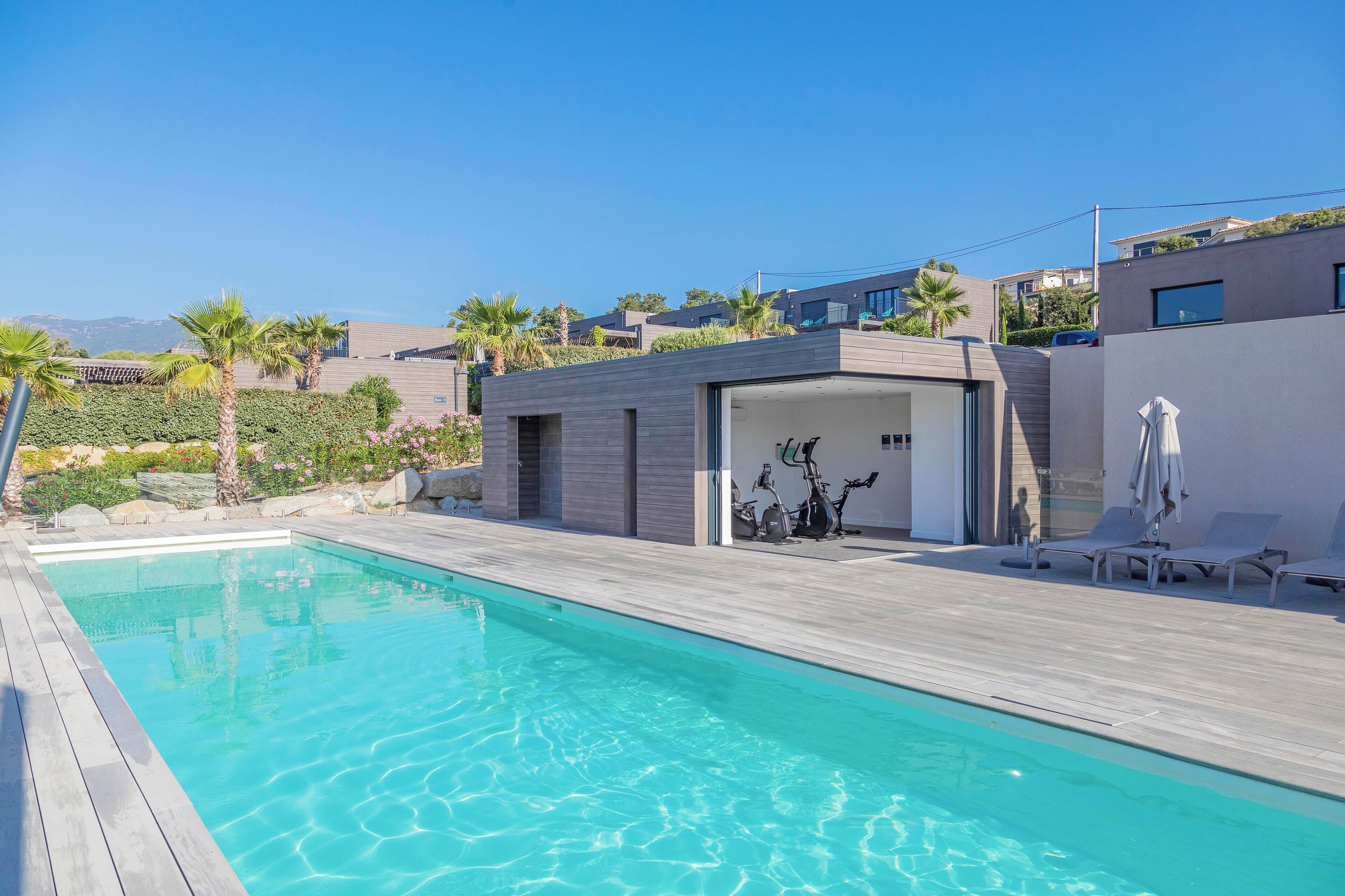 A modern pool area with a wooden deck, a covered exercise area, and a view of neighboring houses.