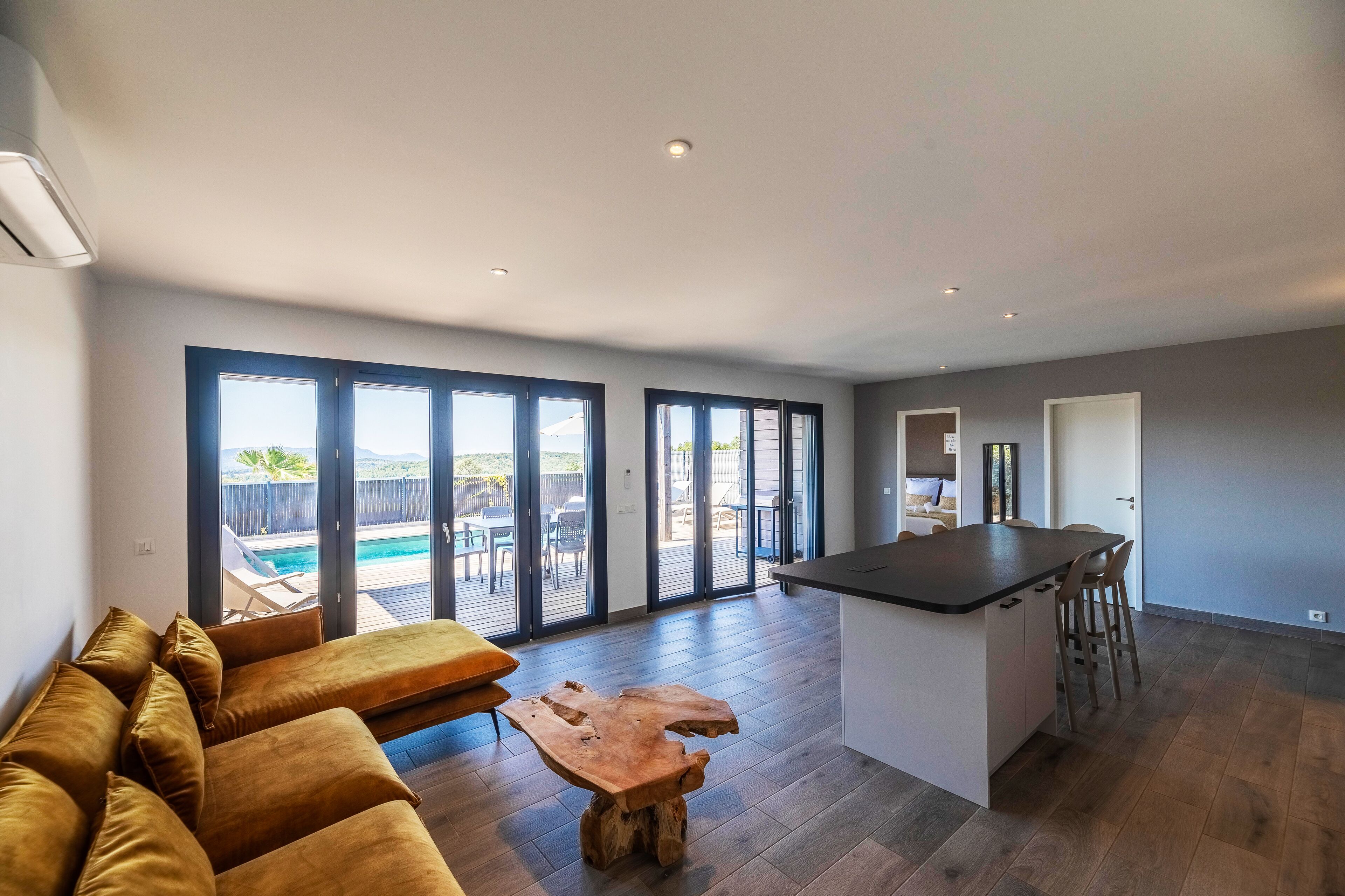 A modern living room with a sofa, a wooden coffee table, and a kitchen area with a bar. Large windows provide a view of a pool and outdoor furniture.