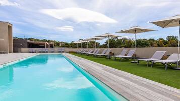 Piscina con sillones de descanso y sombrillas, rodeada por una terraza de madera y un área verde.