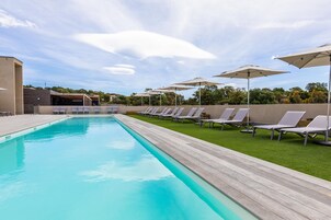 Piscina con sillones de descanso y sombrillas, rodeada por una terraza de madera y un área verde.