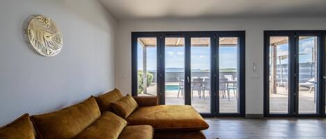 A modern living room with a sectional sofa, a wooden coffee table, and large sliding glass doors leading to an outdoor patio with a pool and furniture.