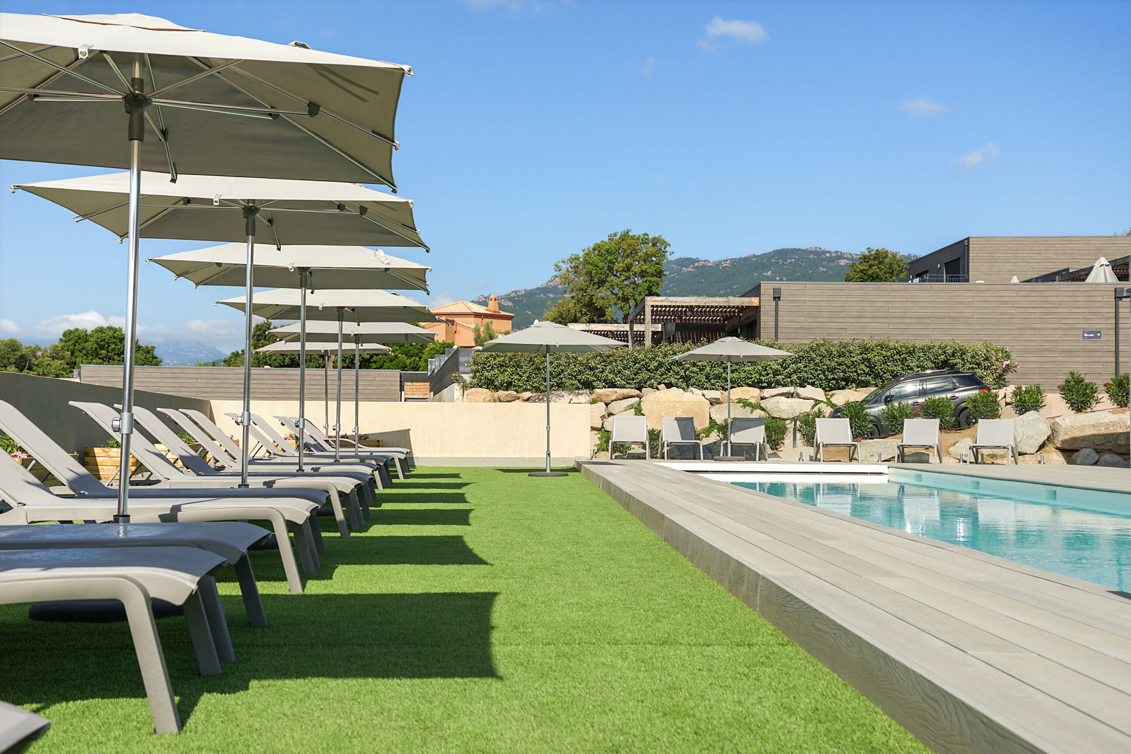 A poolside area with multiple lounge chairs and umbrellas, a swimming pool, and a view of mountains in the background.