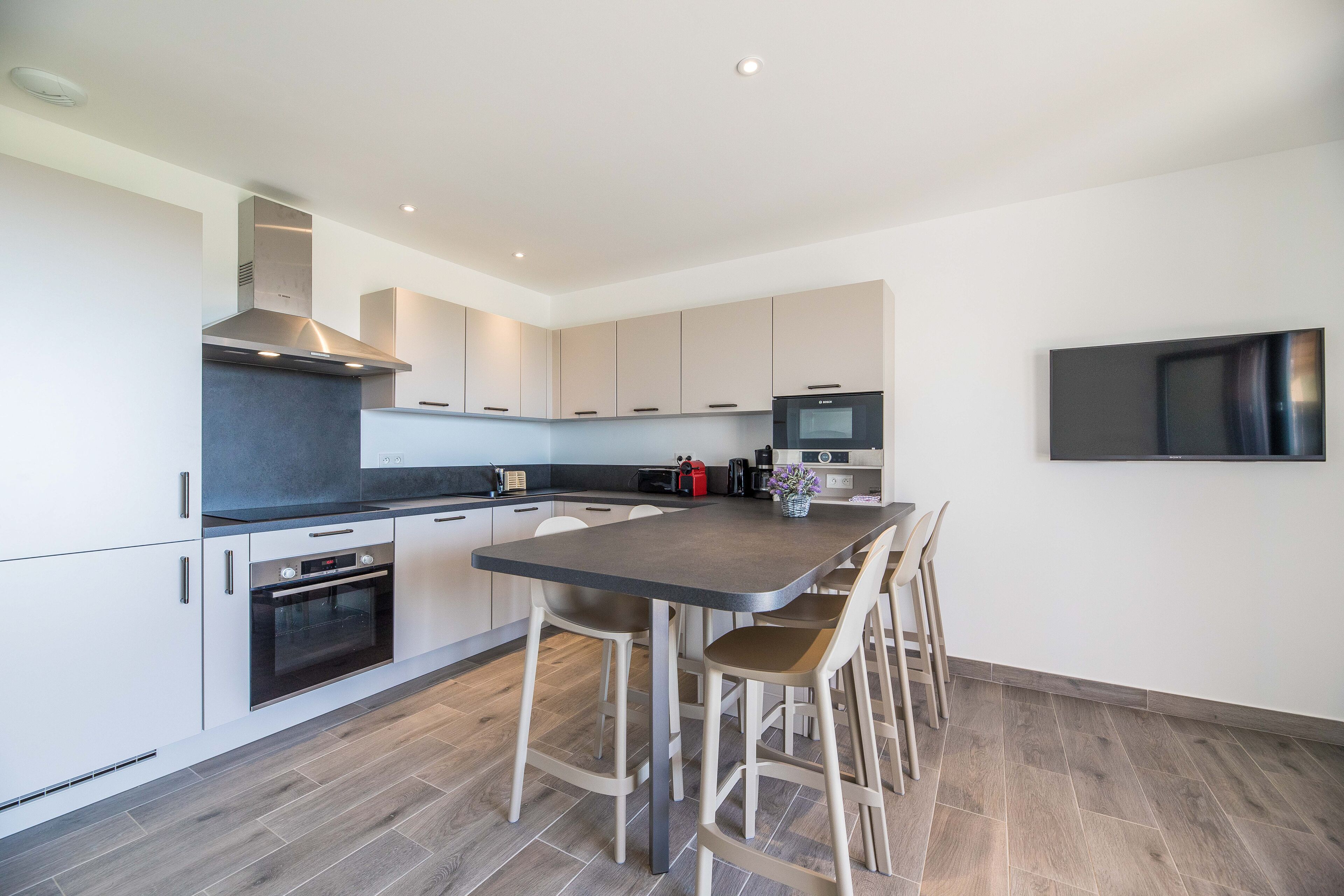 A modern kitchen with a central island, stainless steel appliances, and a flat-screen TV mounted on the wall.