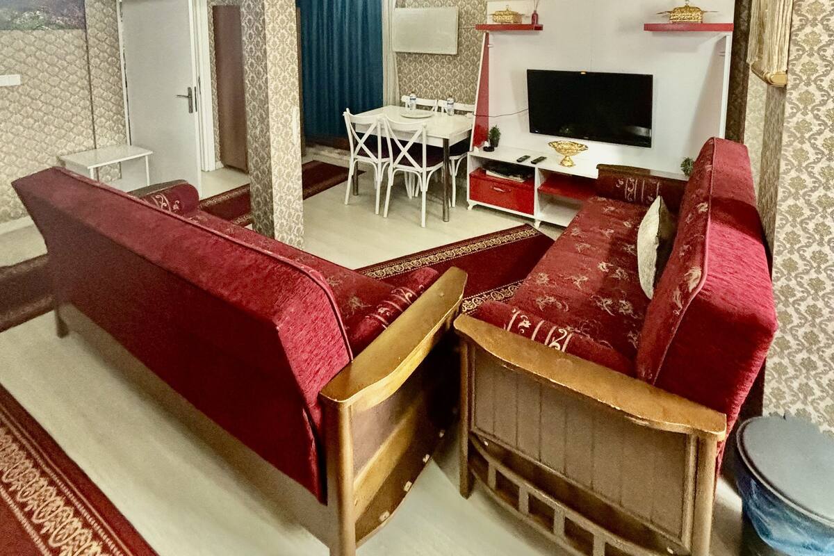 A living room with a red sofa, a wooden coffee table, and a TV. There is a dining area with a white table and chairs.