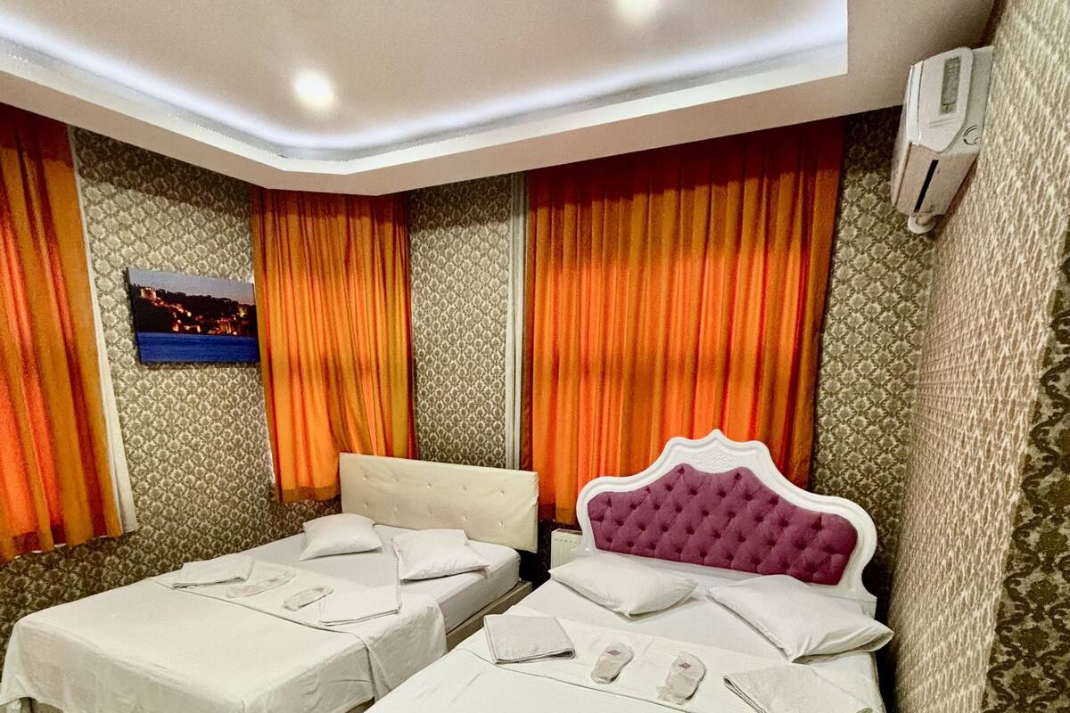 A hotel room with two beds, a large window with orange curtains, a wall-mounted air conditioner, and a painting on the wall.