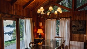 A wooden cabin with a glass dining table, chairs, a chandelier, and a view of a lake. - Beatiful Cabin in Bariloche (San Carlos de Bariloche)