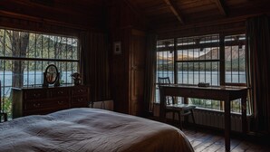 A wooden cabin bedroom with a large bed, a dresser, and a desk by a window overlooking a lake. - Beatiful Cabin in Bariloche (San Carlos de Bariloche)