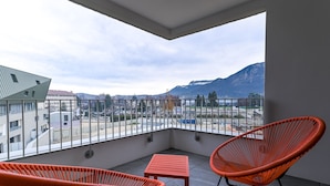 Un balcón con dos sillones de mimbre naranja y una mesita, con vista a un paisaje urbano que incluye edificios y montañas.
