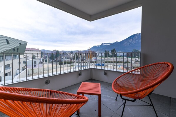 Un balcón con dos sillones de mimbre naranja y una mesita, con vista a un paisaje urbano que incluye edificios y montañas.