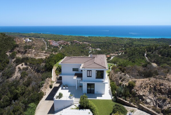 Una casa moderna de dos pisos con techo plano, rodeada de vegetación y vista al mar.