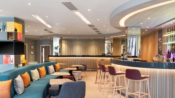 A modern bar area with a curved counter, seating with bar stools, and a sofa with cushions. - Hampton by Hilton London Ealing (London)