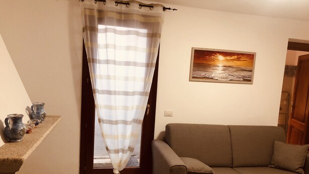A living room with a sofa, a window with curtains, a painting on the wall, and a shelf with decorative items. - Fantastico Budoni Tauanella ARGO02 (Budoni)