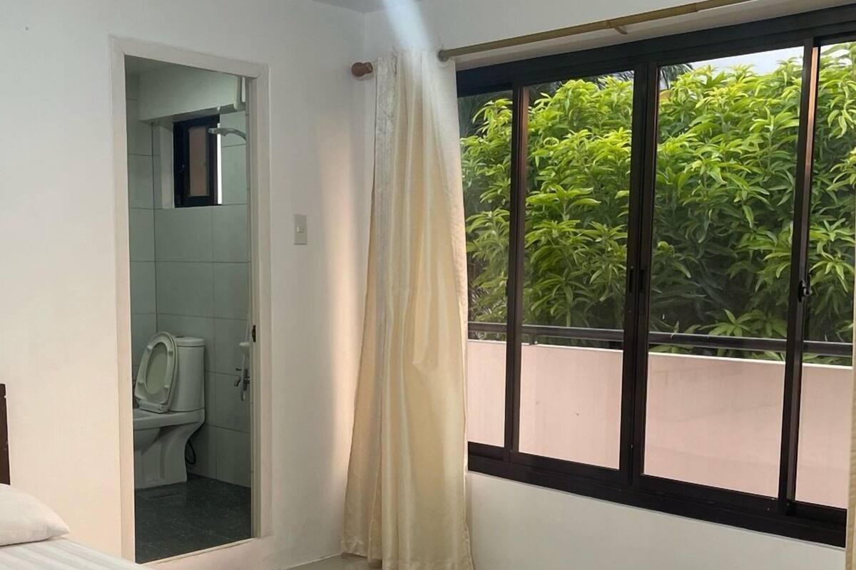 A bedroom with a bed, a window with a view of greenery, a bathroom with a toilet, and a shower.