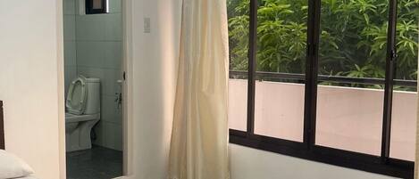 A bedroom with a bed, a window with a view of greenery, a bathroom with a toilet, and a shower.