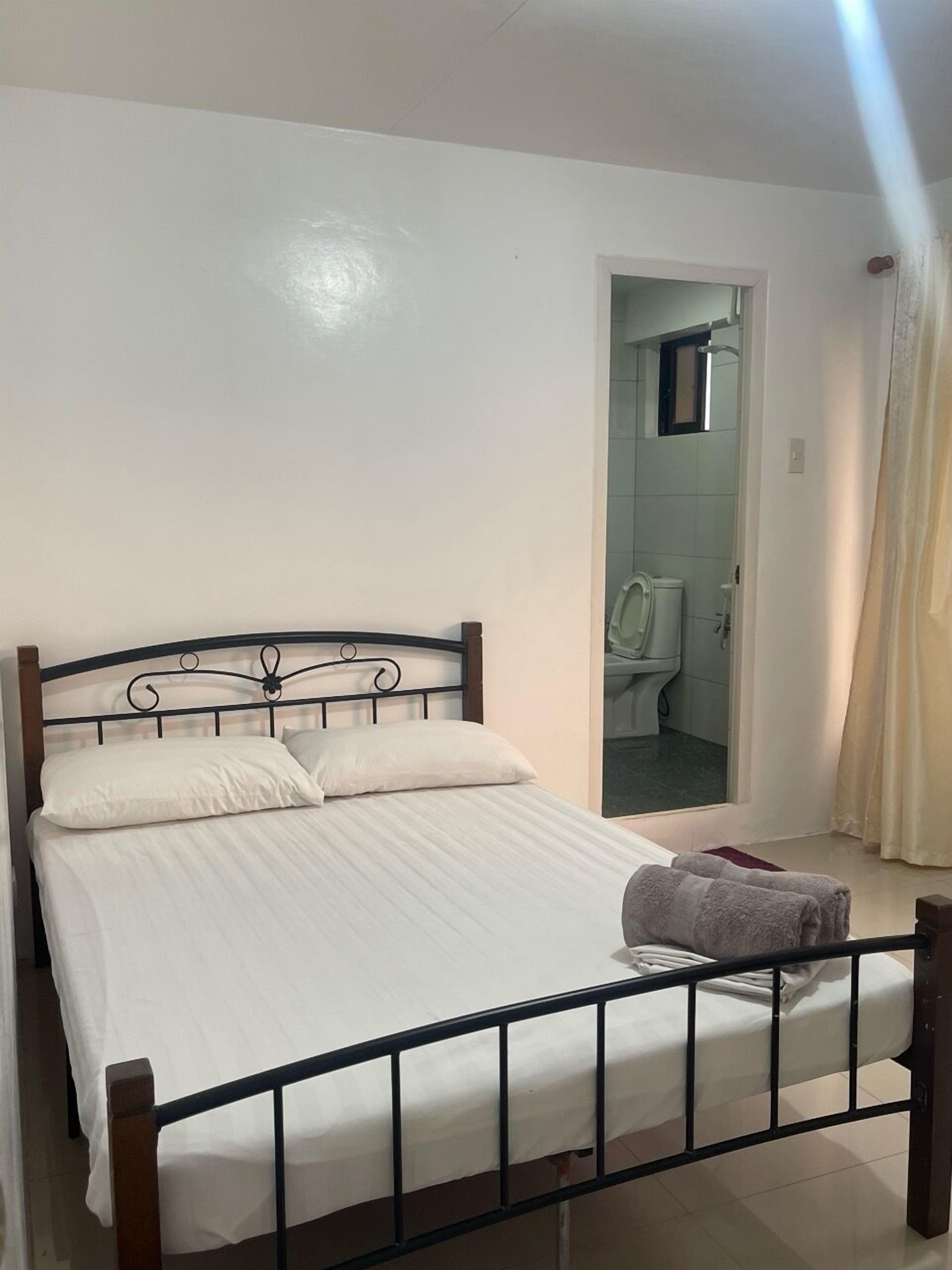 A single bed with a metal frame, a white mattress, and a pillow. There is a towel on the bed. A bathroom is visible through an open doorway.