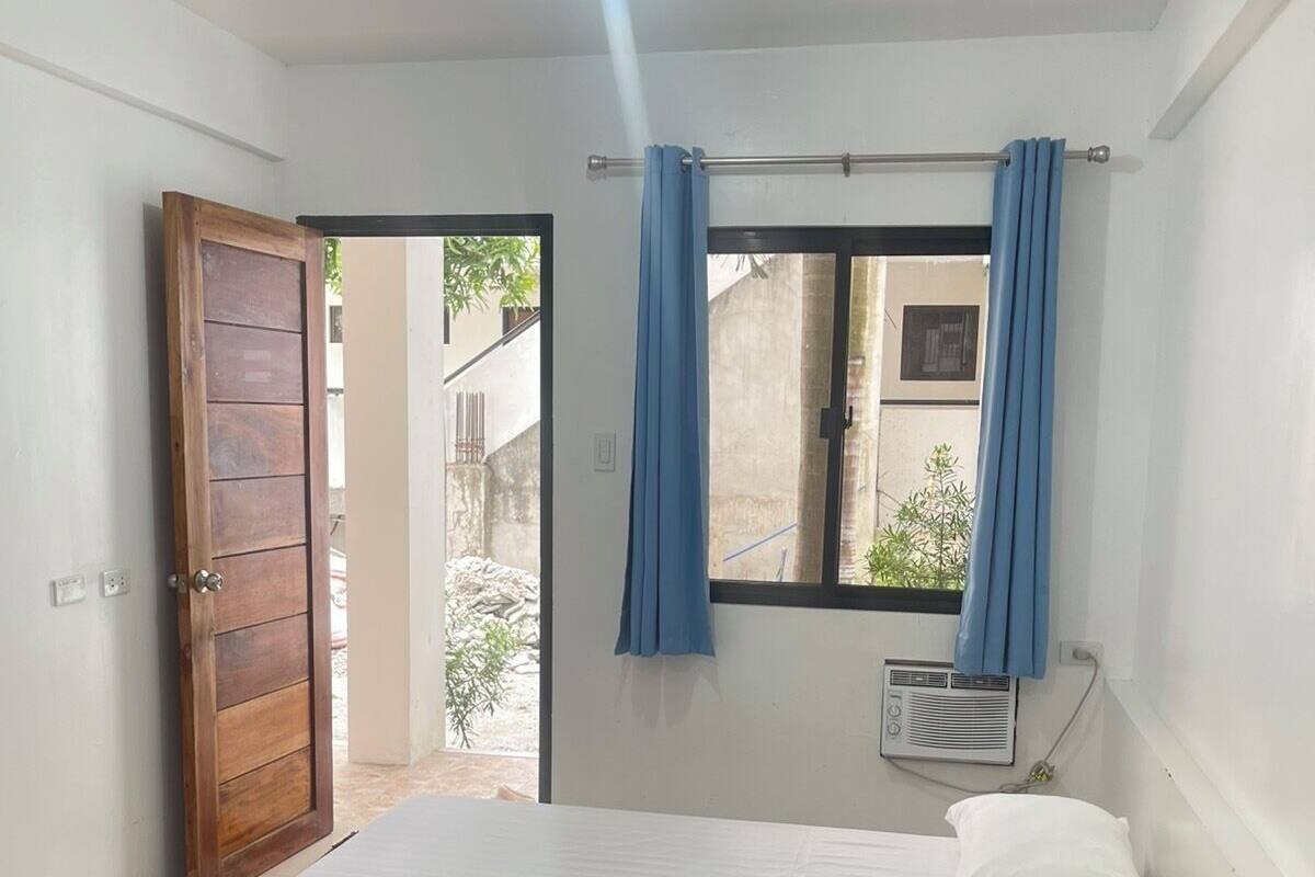 A single bed with a white mattress and a grey towel, a ceiling fan, a window with blue curtains, and a door leading outside.