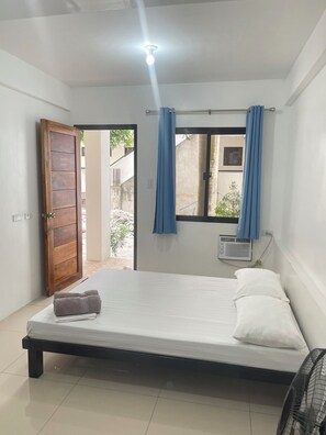 Una cama individual con colchón blanco y toalla gris, un ventilador de techo, una ventana con cortinas azules y una puerta que da al exterior.