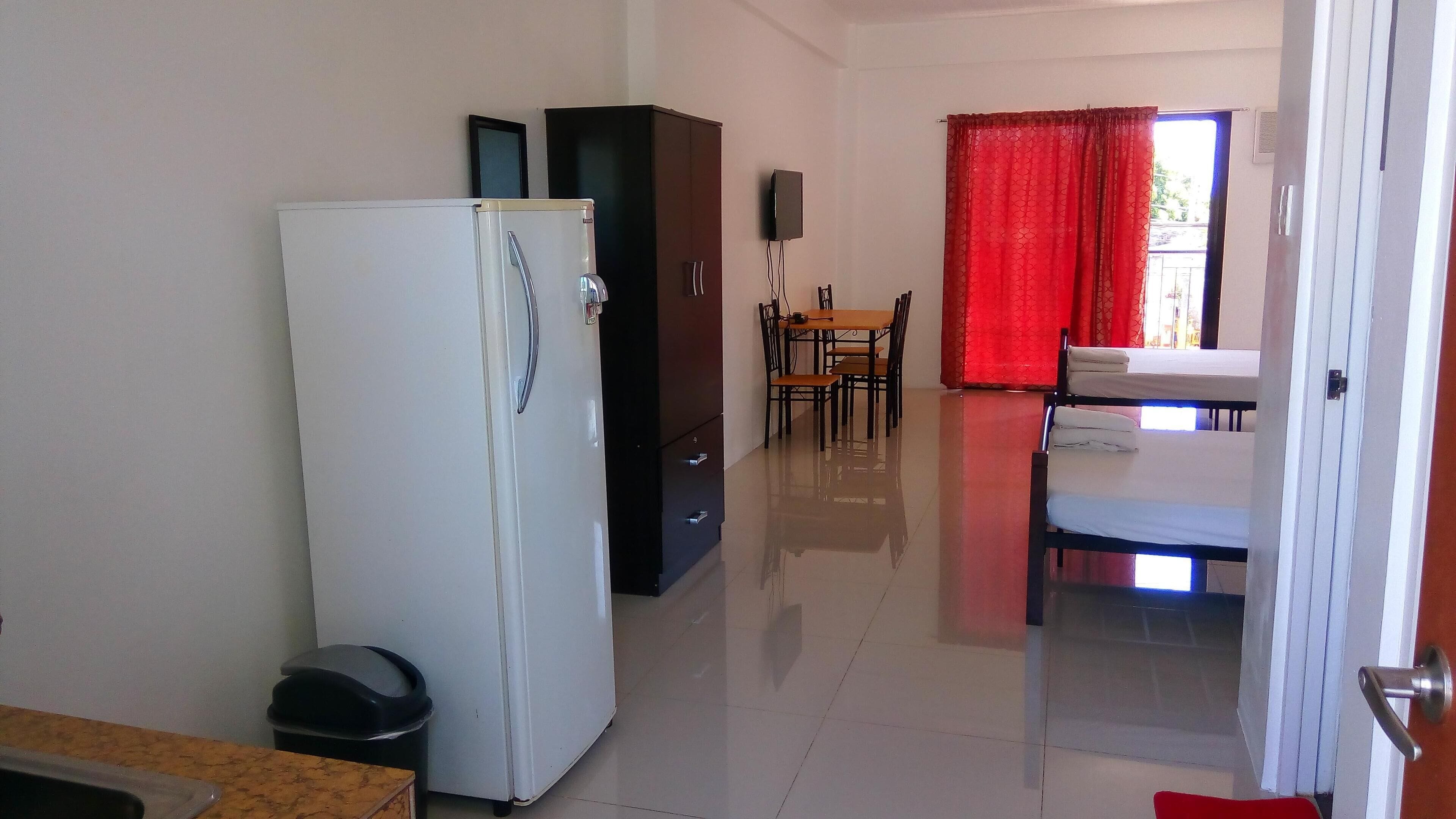 Habitación de hotel compacta con un refrigerador blanco, una pequeña mesa con una silla, una mesa de comedor con sillas y una cortina roja.
