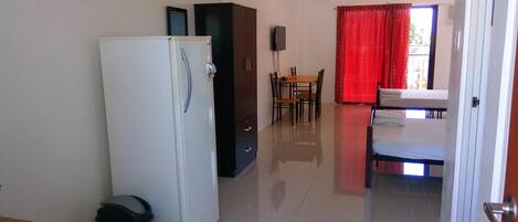 Habitación de hotel compacta con un refrigerador blanco, una pequeña mesa con una silla, una mesa de comedor con sillas y una cortina roja.