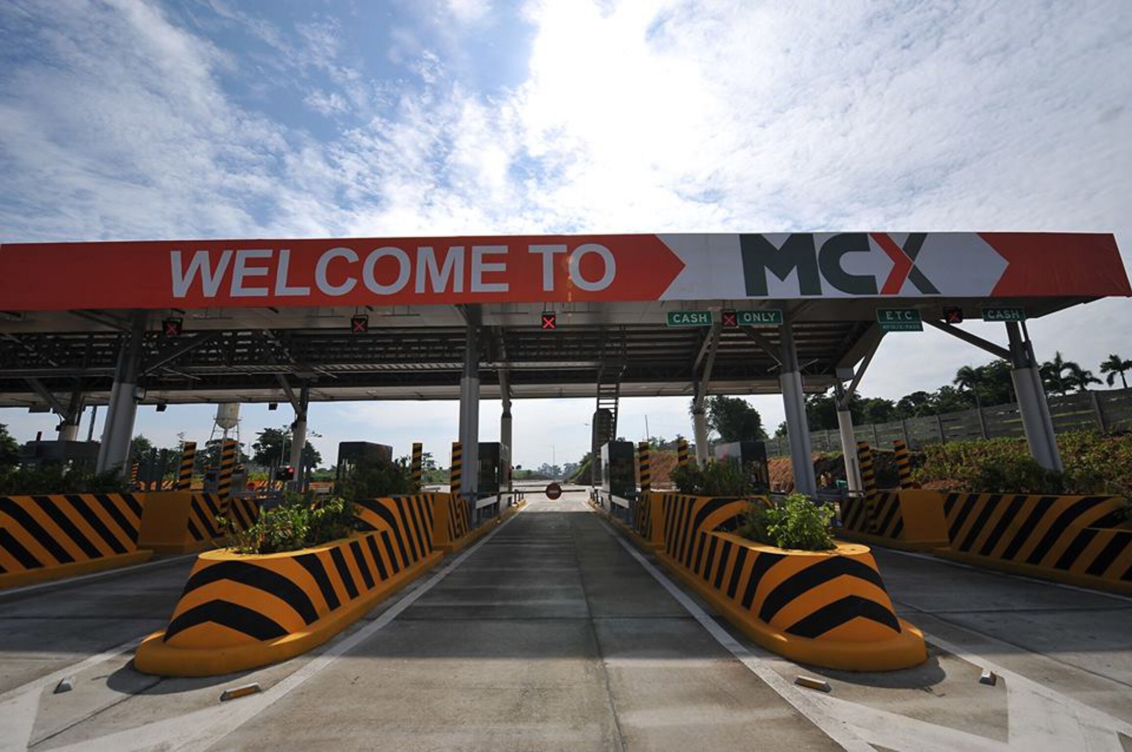 A toll booth entrance with a 'WELCOME TO MCX' sign, cash and ETC payment options, and a clear sky.