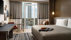 A hotel room with a bed, a desk, a chair, a small table, and a balcony with a view of high-rise buildings.