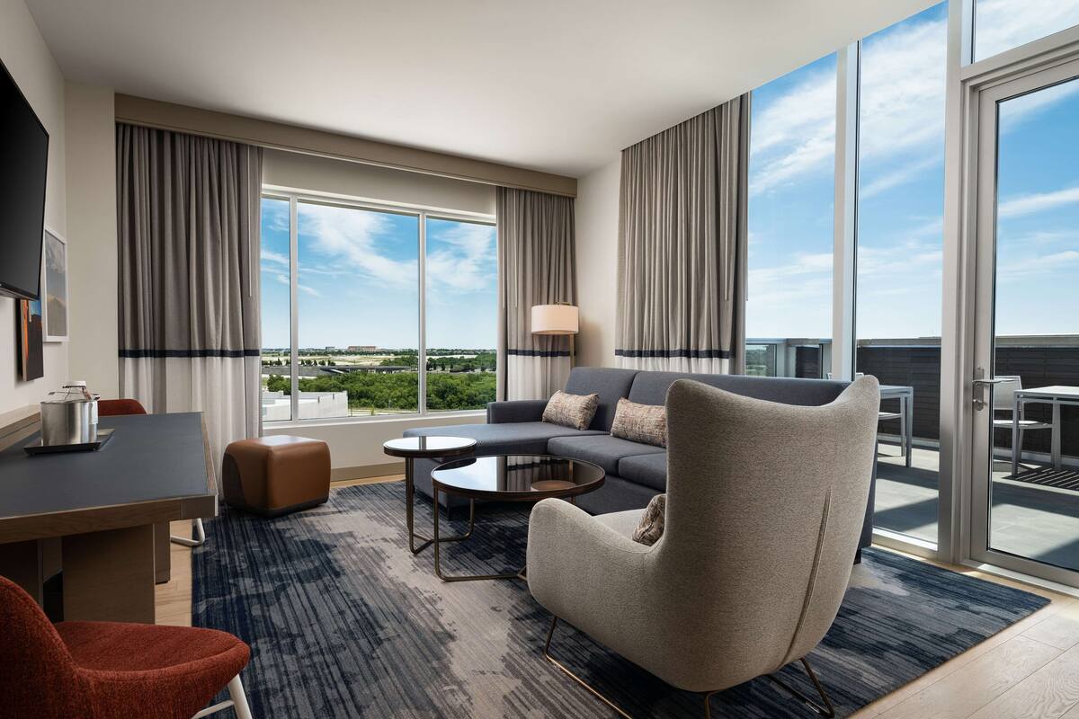 A modern hotel room with a large window, a sofa, a coffee table, a chair, and a desk with a lamp.